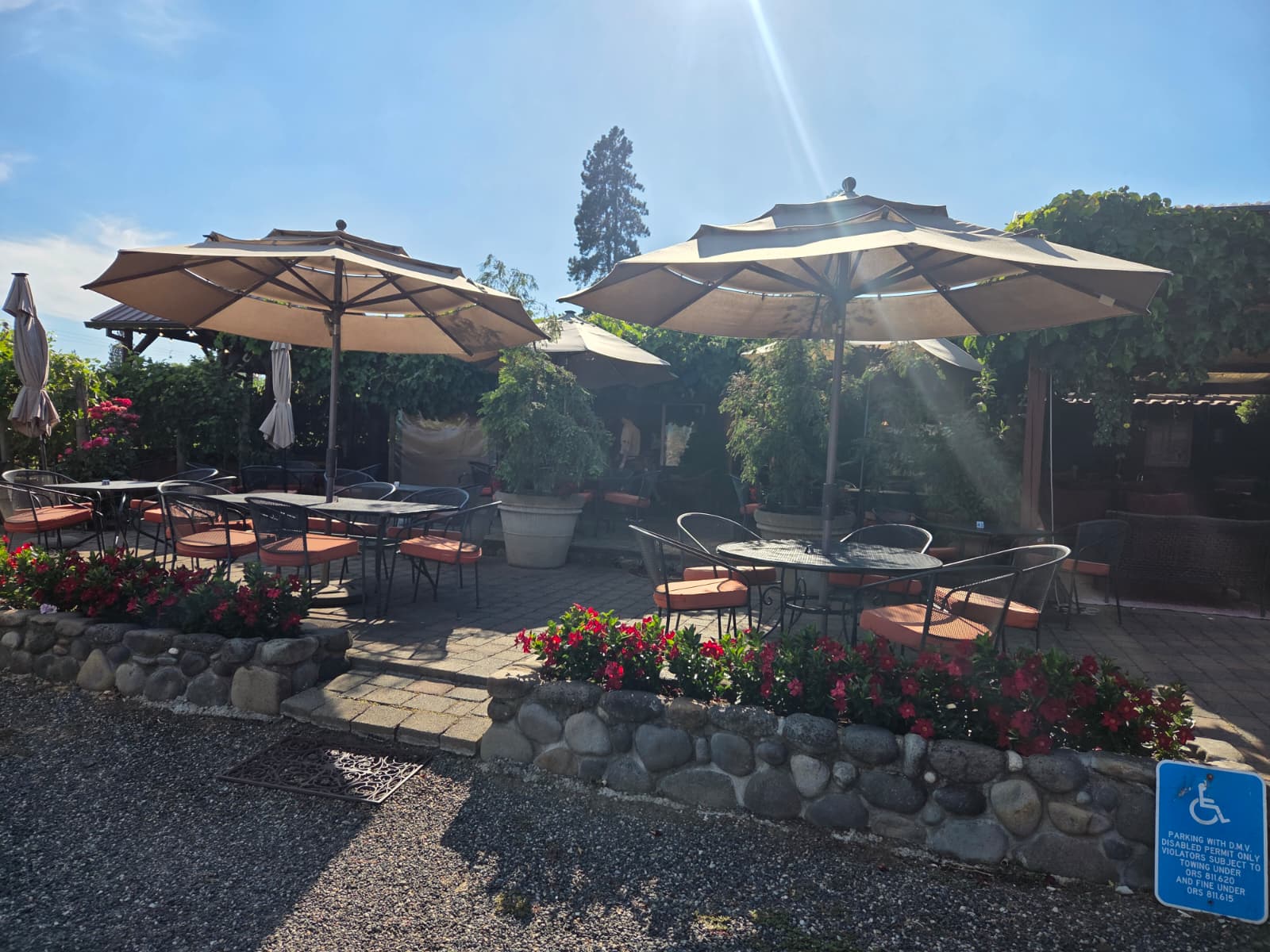 Sunlit outdoor patio at Marchesi Vineyards with tan umbrellas, wrought-iron tables and red flowers along a river-rock wall