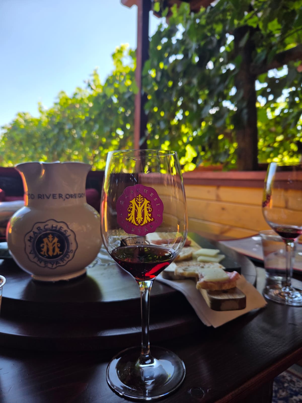 Glass of red wine with the Marchesi Vineyards logo beside a Hood River, Oregon ceramic pitcher and a charcuterie board overlooking the vines