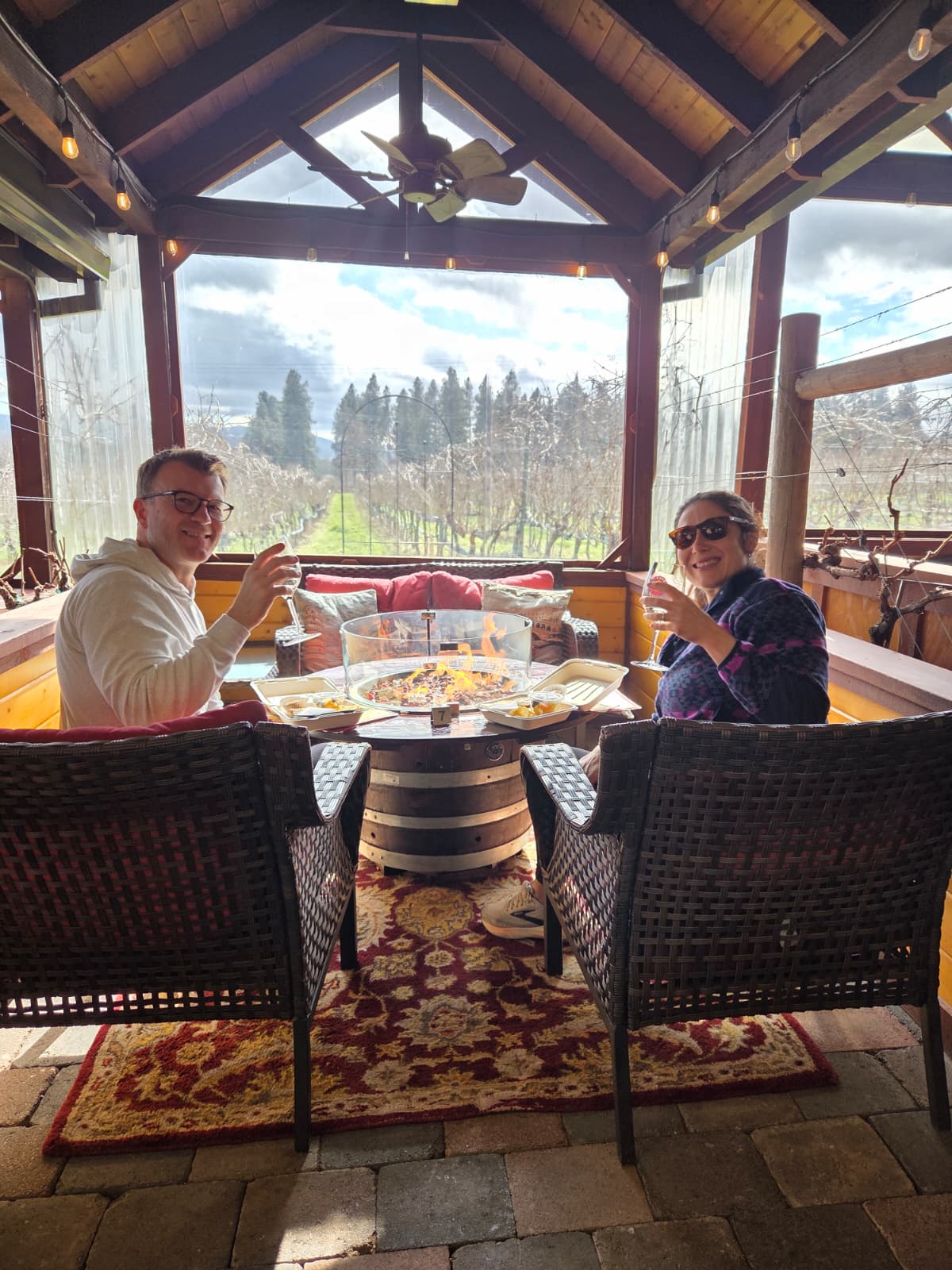 A couple raising wine glasses at a barrel table with a fire pit inside a covered vineyard gazebo, dormant vines and mountains beyond